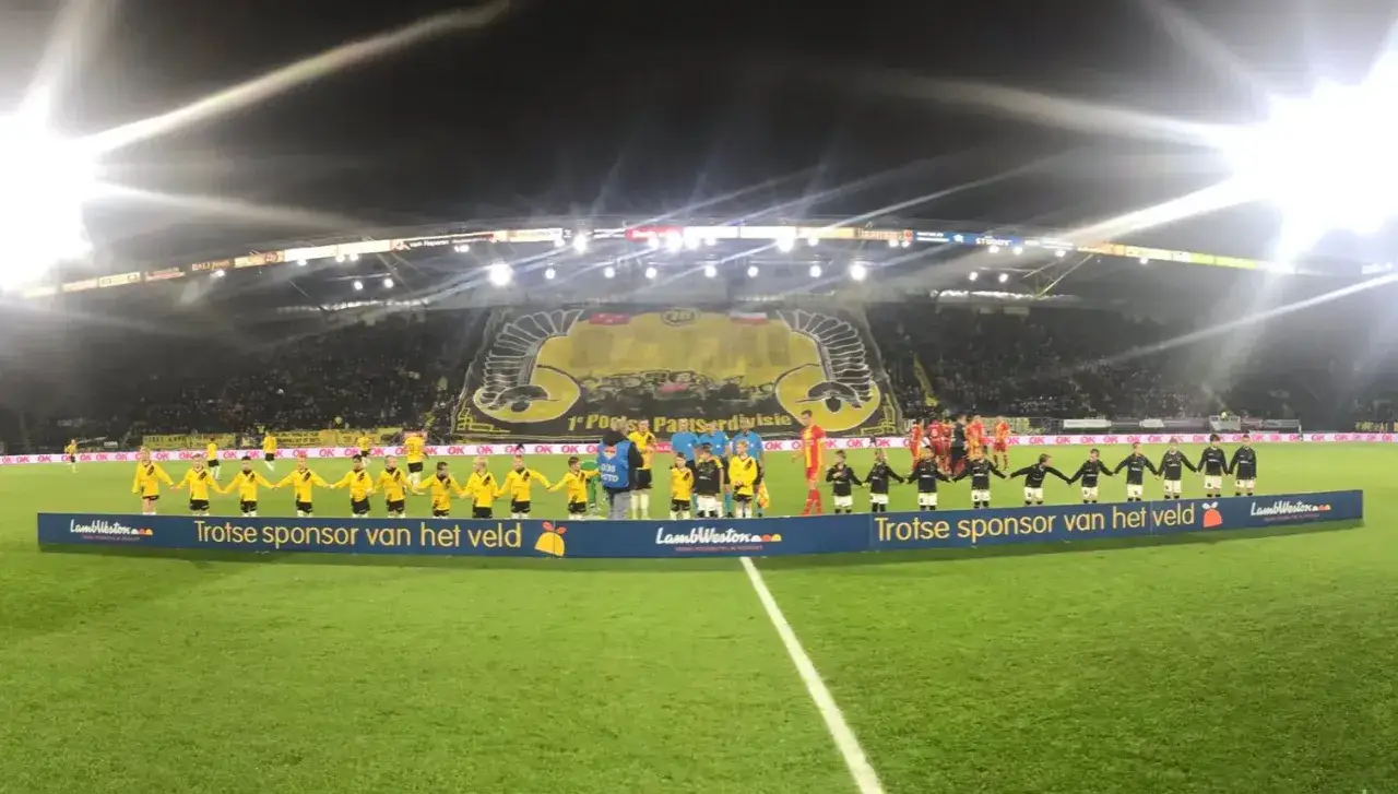Dzieci w żółtych strojach stoją w rzędzie, trzymając się za ręce na boisku NAC Breda. Na trybunach tłum kibiców.
