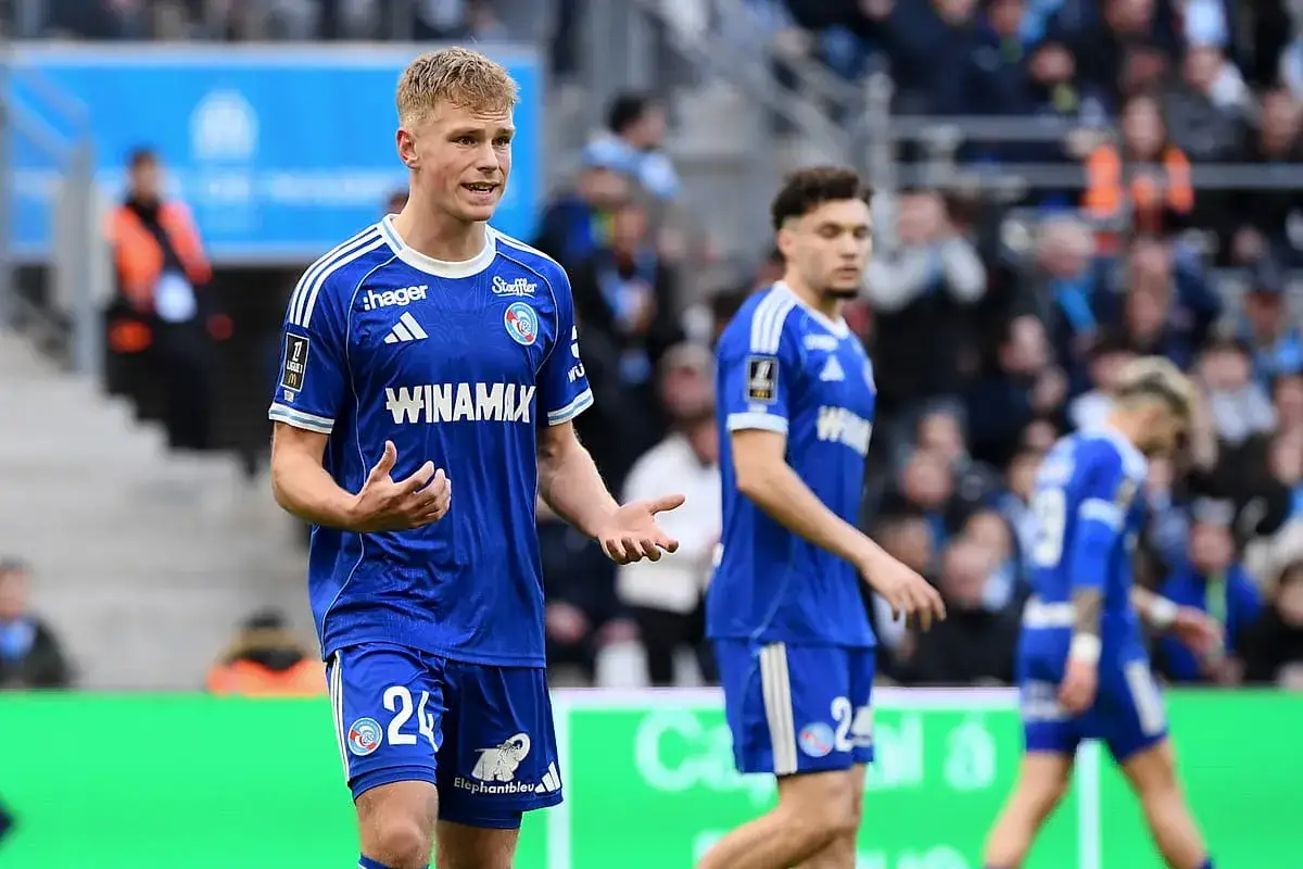 Piłkarz RC Strasbourg w niebieskiej koszulce z numerem 24, gestykuluje podczas meczu. W tle inni zawodnicy i kibice.