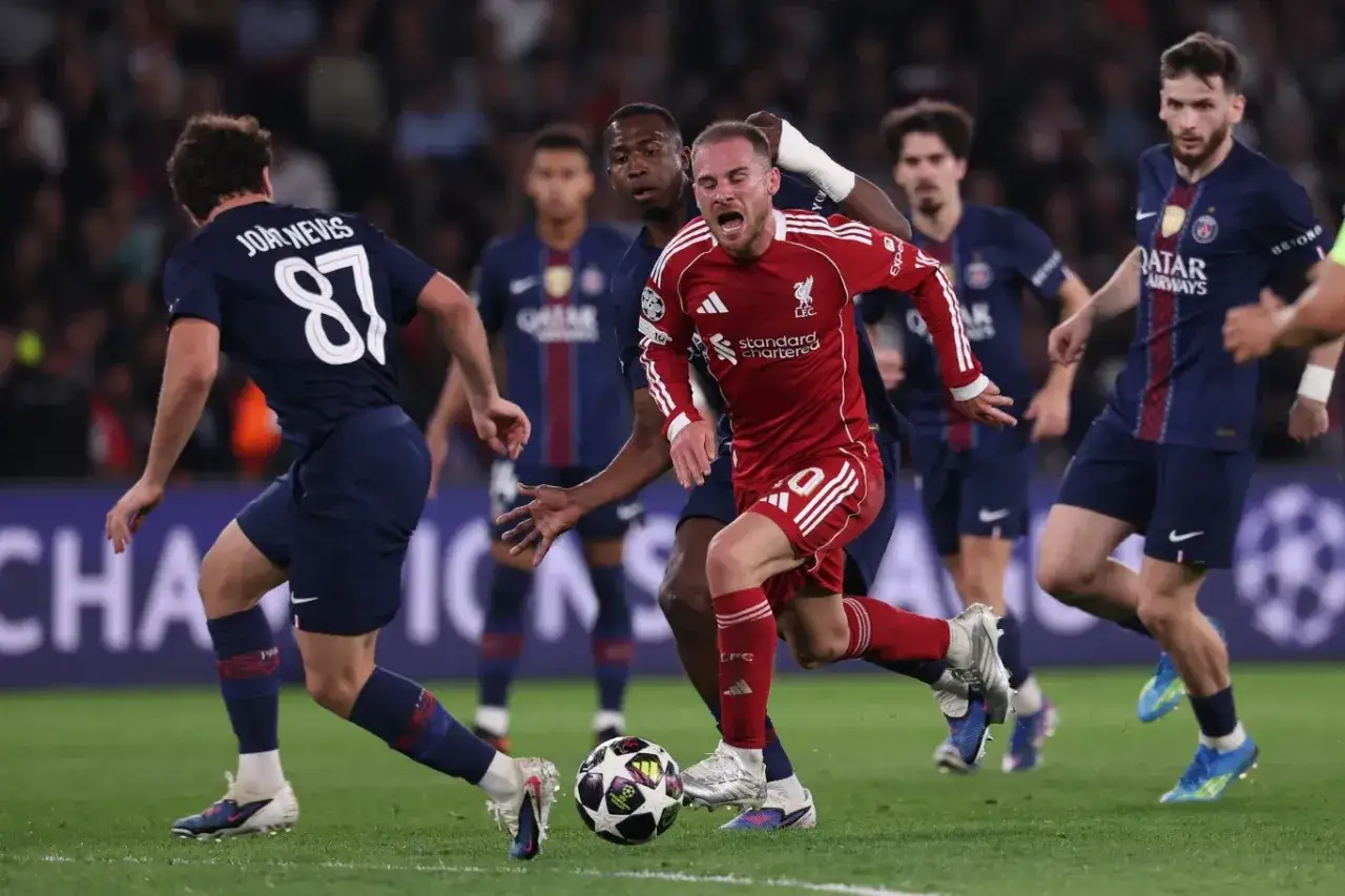 Intensywna akcja na boisku podczas meczu PSG – Liverpool. Piłkarz w czerwonej koszulce Liverpoolu walczy o piłkę z zawodnikami PSG.