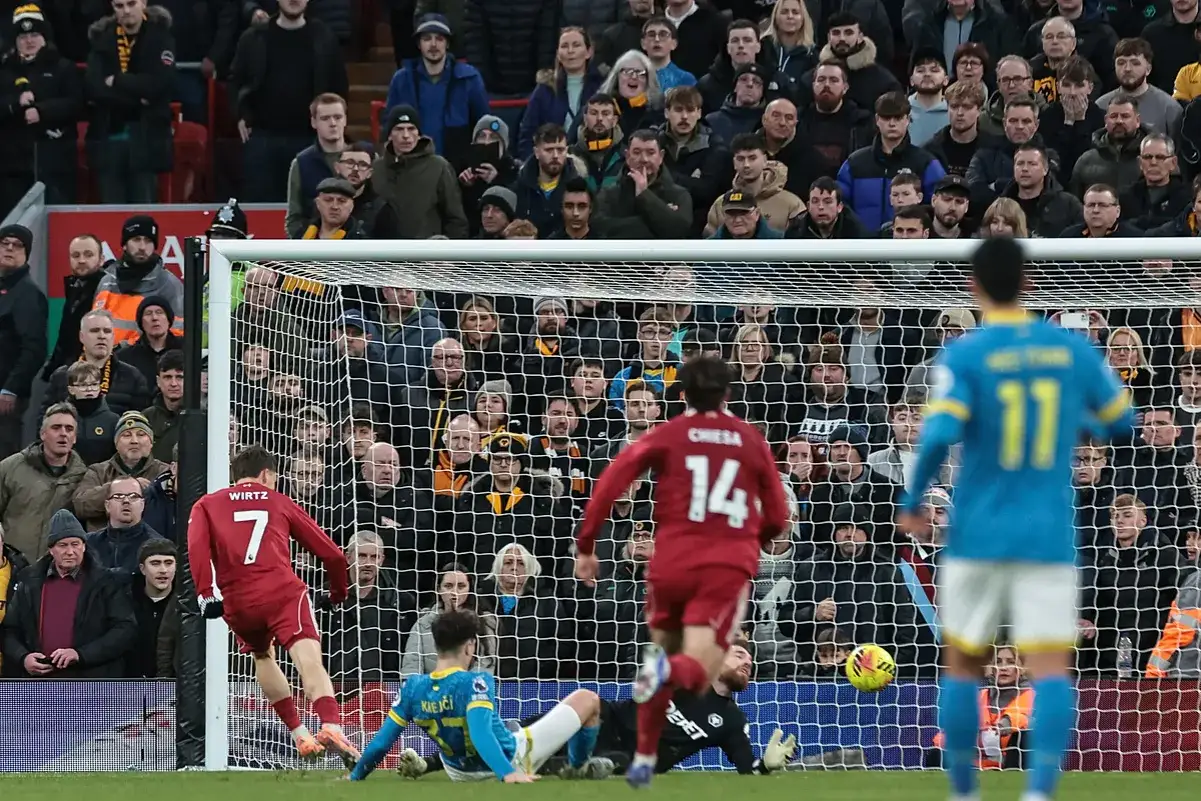 Piłkarz w czerwonej koszulce z numerem 7 strzela gola w meczu Leeds – Liverpool. Bramkarz w niebieskim stroju próbuje interweniować.