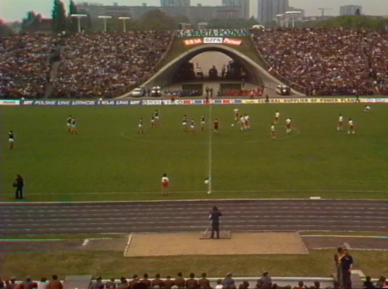 Mecz piłkarski na stadionie KS Warta Poznań. Pełne trybuny kibiców, zawodnicy na murawie.