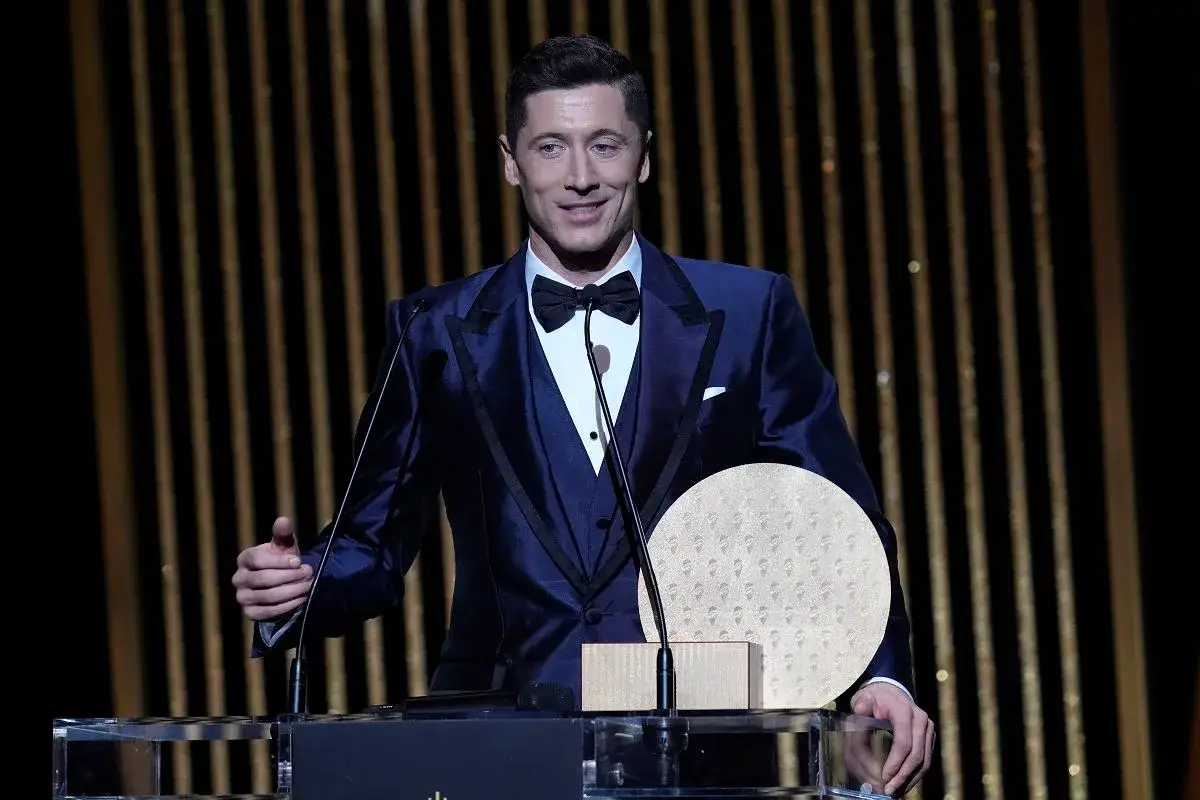 Robert Lewandowski z dumą prezentuje swoją złotą piłkę zwycięzcy, ubraną w elegancki, granatowy garnitur.