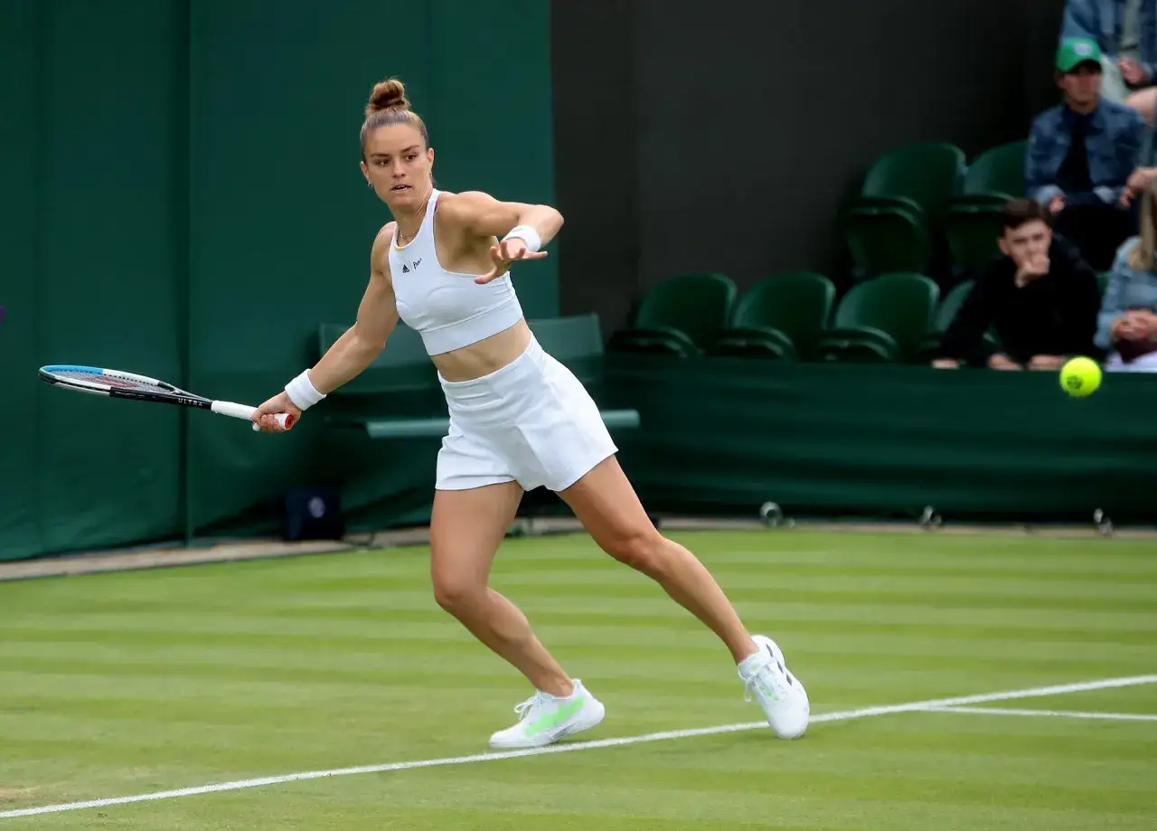 Maria Sakkari w akcji na trawiastym korcie, w białym stroju tenisowym, z rakietą w dłoni.