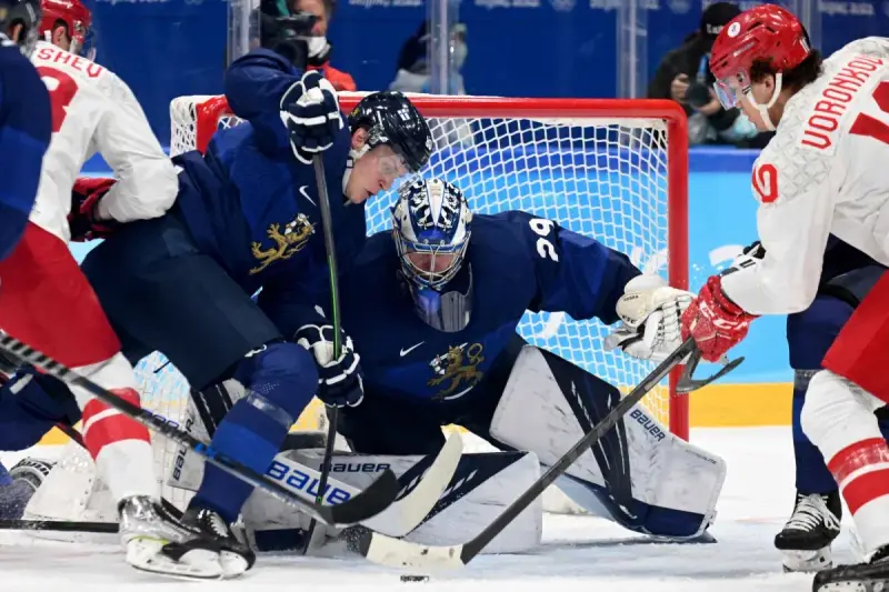 Bramkarz Finlandii broni strzał. W tle zawodnicy walczą o krążek, dążąc do zwycięstwa, jak przystało na mistrzów olimpijskich w hokeju na lodzie.